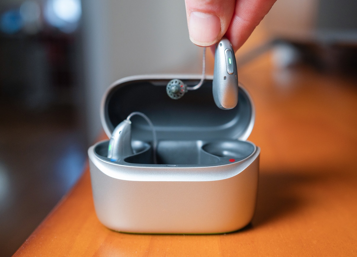 Hand holding hearing aid over charging case on table.